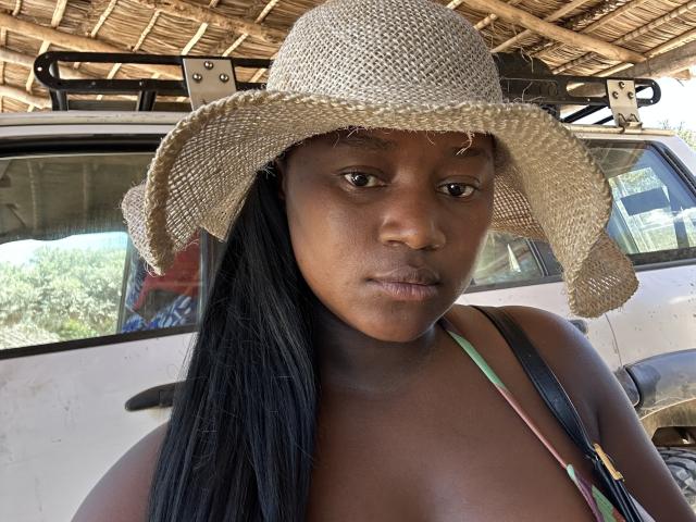 PassionFrutz, une femme à la peau foncée, portant un chapeau de paille, avec des cheveux noirs, une bretelle verte et noire, avec une expression sérieuse, un camion blanc en arrière-plan, un toit en bambou, ensoleillement, de légères marques de bronzage visibles..