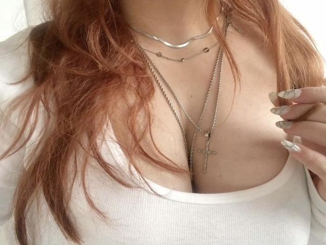 El pecho de GoldenGinger, mostrando sus grandes senos en una camiseta blanca con textura, con cabello largo y rojo, múltiples collares de plata, y su mano con uñas cuidadas cerca de su escote..