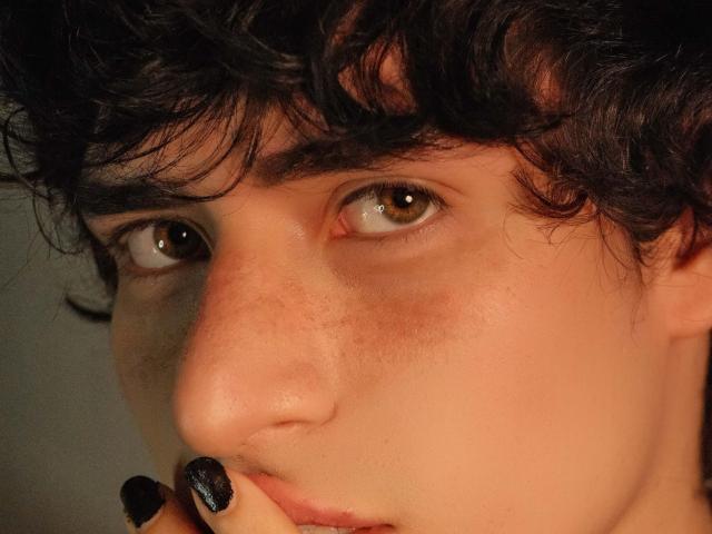 DemonFox a les cheveux noirs bouclés, les yeux marron et porte du vernis à ongles noir. Un doigt est près de ses lèvres légèrement entrouvertes, révélant une expression subtile et séduisante..