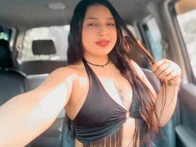 JadeRouxx with long black hair, wearing a black halter top with fringe, holding her hair, showing her tits, and a tattoo on her chest, in a car.