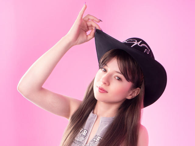 VioletaNova with long brown hair, wearing a black hat, lifting it with her right hand, pink background, gray sleeveless top, subtle makeup, gentle smile, pink nails, looking at camera.