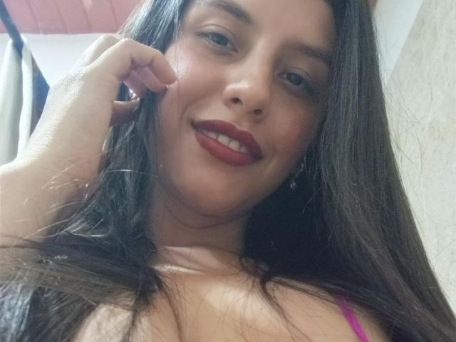 MadissonFontaine with long brown hair, red lipstick, and pink bra, smiling at the camera, showing her tits, hand touching her cheek. Her skin is light brown.