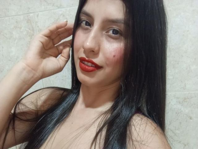 MadissonFontaine has long black hair, red lipstick, and a small zit on her right cheek. She's smiling, touching her ear with her right hand. Her tits are visible.