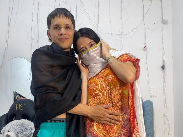 AnjaliRohan with short brown hair, black shawl, and teal underwear, hugs a woman in an orange and red patterned sari, white mask, and yellow forehead mark.