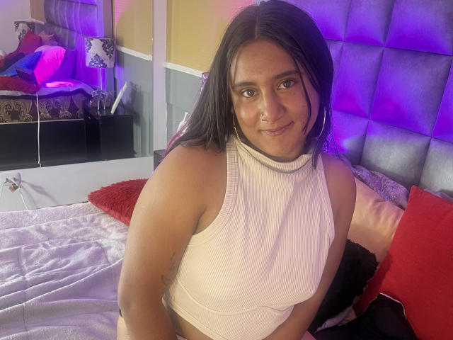 MarianDiore, avec une peau marron clair et des cheveux noirs, portant un crop top blanc côtelé, montrant de petites seins, assise sur un lit avec des coussins rouges et gris..
