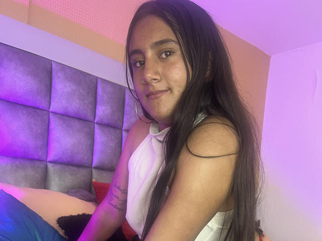 MarianDiore, avec de longs cheveux noirs, un anneau au nez, un haut blanc à encolure dégagée, un tatouage sur le bras droit, assise sur un lit, souriant à la caméra, éclairage violet et rose, cuisses visibles..