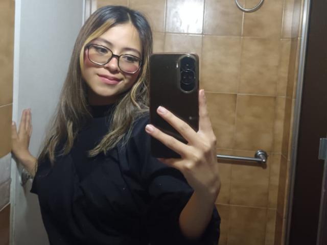 AnittaRouses with glasses, smiling, holding a phone, black shirt, brown tile bathroom, left hand on wall, right hand holding phone, blonde hair with brown highlights, cute.