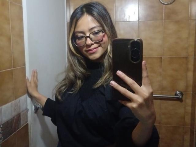 AnittaRouses with glasses, black turtleneck, and long brown hair, holding a phone, smiling, showing tits, against tiled wall, hand on door, brown tiles in background.