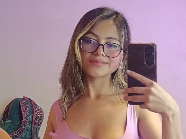 AnittaRouses with glasses, pink tank top, and blonde-streaked brown hair, taking a selfie in a mirror, showing small tits, holding a black phone, smiling slightly.