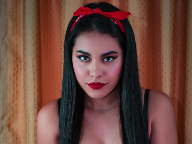 PaolaYKatty with long black hair, red bandana, red lipstick, and bold eye makeup, looking directly at the camera, wearing a black top, with a silver necklace.