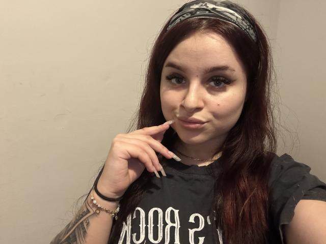 LucieRed with long red hair, gray bandana, black shirt, tattooed arm, finger to lips, black nails, bracelet, subtle smile, light skin, makeup, and a cute, sexy look.
