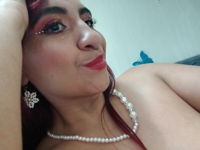 NahelaBigMelonsX with red lipstick, pearl necklace, and red eyeliner, wearing a star earring, lying down, looking up with a slight smile, hand on head.