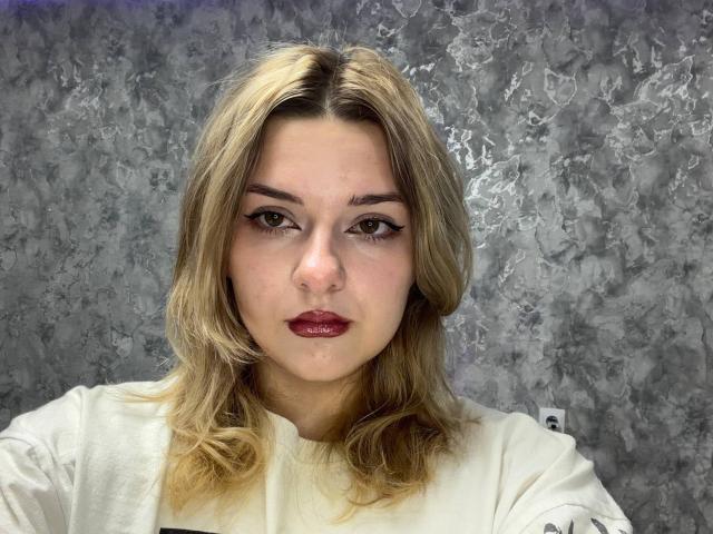 AdrianePittillo has blonde hair, light skin, and red lipstick, wearing a white shirt, with a slightly bored expression and subtle eye makeup, against a textured grey background.