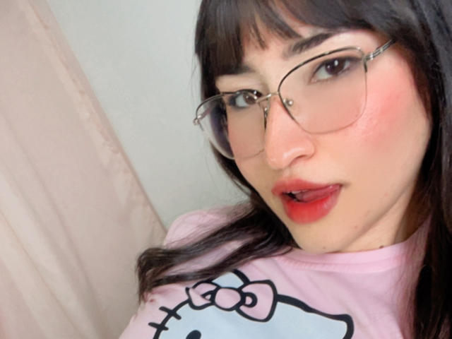 JackyBell con gli occhiali, labbra rosa, rossetto rosso e capelli neri, indossa una maglietta rosa di Hello Kitty, guarda la fotocamera con un'espressione di bocca leggermente aperta..