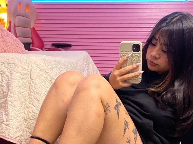 AmaraKazumi with black hair and tattoos on her thigh, taking a selfie in a pink room, wearing a black top, legs crossed, holding a gold phone, slightly pouting.