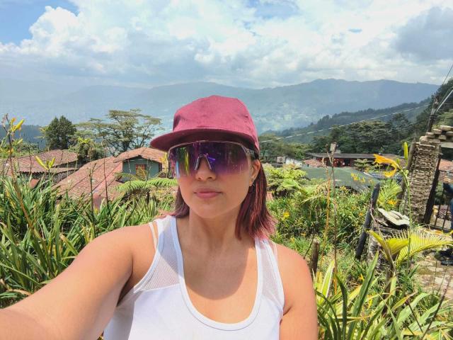 FetishWayraa with red cap, purple sunglasses, and white tank top, standing in front of green foliage, showing her small tits, and smiling at the camera.