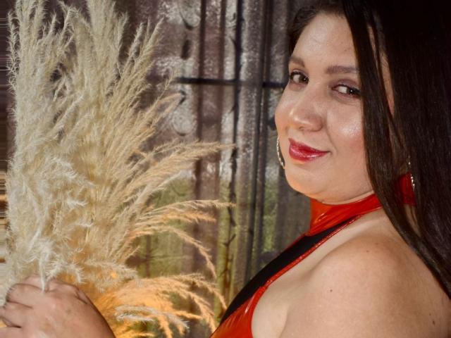 PaulinaStorm with shiny red halter top, holding fluffy beige pampas grass, smiling at camera, brown hair, red lipstick, hoop earrings, fair skin, soft light, slight side profile.