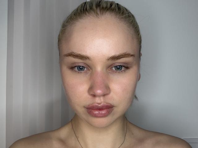 LeannaLubrano's Gesicht, mit blasser Haut, blauen Augen, blondem, zurückgebundenem Haar, Augenbrauen und rosafarbenen Lippen, trägt eine einfache goldene Halskette und blickt direkt in die Kamera..