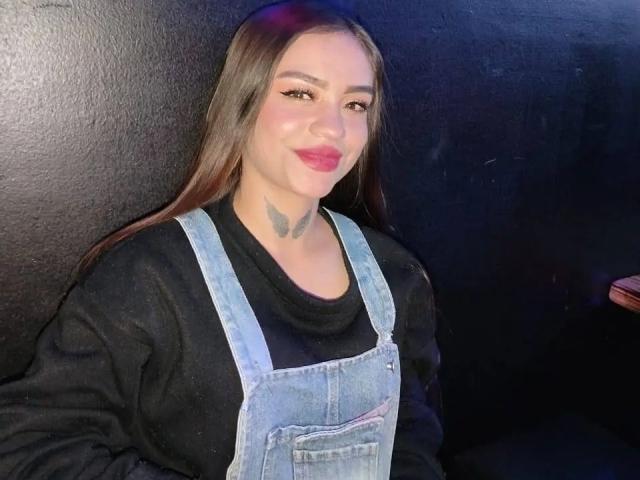 AnnaLadera with long brown hair, black shirt, blue denim overalls, black butterfly tattoo on neck, pink lipstick, smiling, black textured wall in background, right hand not visible.