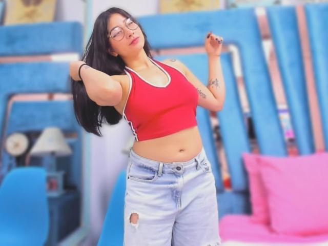 Genesiss has medium tits in a red sports bra, wearing light blue, ripped jeans, and glasses, with long black hair, standing in a bedroom, hands in hair.