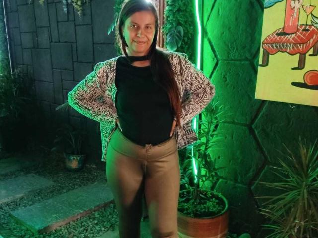 AmbeRouse with hands on hips, wearing a black top, tan pants, and a patterned jacket, standing in front of a green neon light, smiling. Her ass is visible.