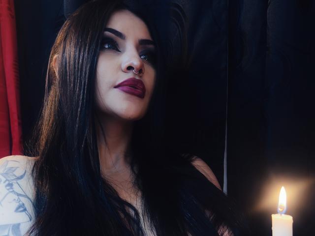 LombanaX with long black hair, dark red lipstick, nose ring, tattooed arm, and serious expression, illuminated by a candle's soft flame. Her tits are partially visible.