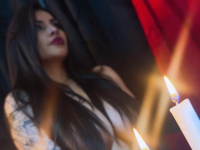 LombanaX with long dark hair, red lipstick, and tattoos, blurred in the background, illuminated by two burning candles in the foreground.