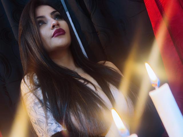 LombanaX with long brown hair, nose ring, and red lipstick, looking up with a slight smile, has visible tattoos on her right arm, and is near two lit candles.