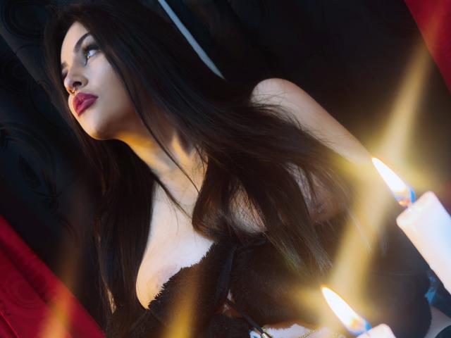 LombanaX with long black hair, red lips, and black lace top, looking up with a serious expression. Two lit candles in the foreground. Boobs visible under lace.