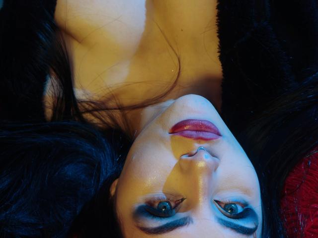 LombanaX's face upside down, blue eyeshadow, red lipstick, black hair, light skin, looking at camera, slightly flushed, wearing black fur, red fabric in background.