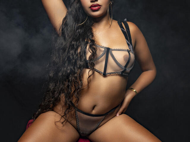 JeimyCampos with long curly black hair, wearing a sheer black bra and panties, showing her boobs and pussy, red lipstick, gold earrings, and a gold bracelet.