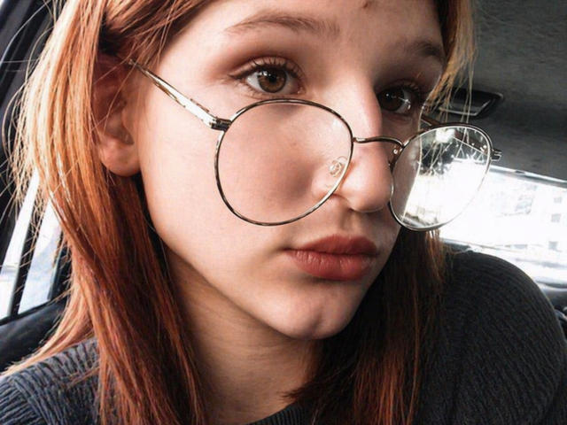 KrystaFessels Gesicht, mit runder Brille und einem leichten Spiegelbild, hat hellbraunes Haar, helle Haut und volle, rosafarbene Lippen. Sie sitzt in einem Auto und blickt direkt in die Kamera..