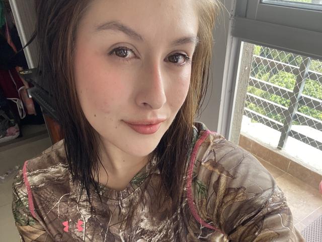 IsabellaPalacio with light skin, brown eyes, and brown hair, wearing a camouflage shirt, has a small mole near her mouth, and is sitting near a window with a chain fence.