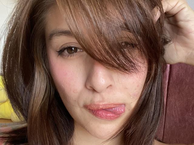 IsabellaPalacio has brown hair with bangs, a pink nose, and is sticking out her pink tongue. She has light skin with a few small blemishes on her face.