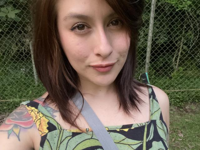 Isabella Palacio ha gli occhi marroni, un naso piccolo e un leggero sorriso. Indossa un abito floreale verde e nero con una cintura e un tatuaggio di una rosa visibile..