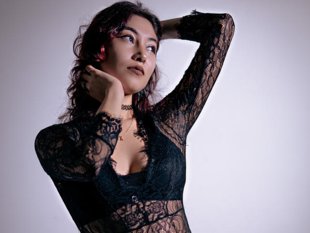 AliCipher, com cabelo preto encaracolado, vestindo uma blusa preta de renda que revela seus pequenos seios, com a mão no pescoço, olhando para o lado, batom vermelho, colar, fundo branco..