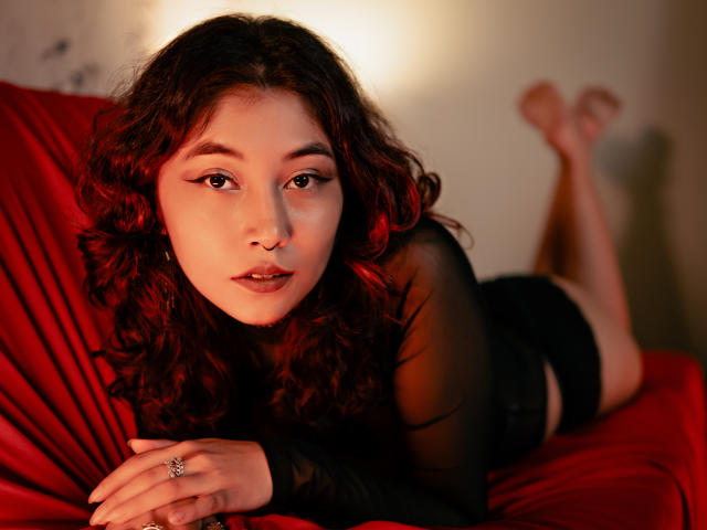 AliCipher, avec ses cheveux roux et noirs bouclés, est allongée sur un lit rouge, vêtue d'un haut noir transparent, de shorts noirs et portant une bague. Ses jambes sont relevées et elle regarde l'appareil photo, avec un éclairage doux..