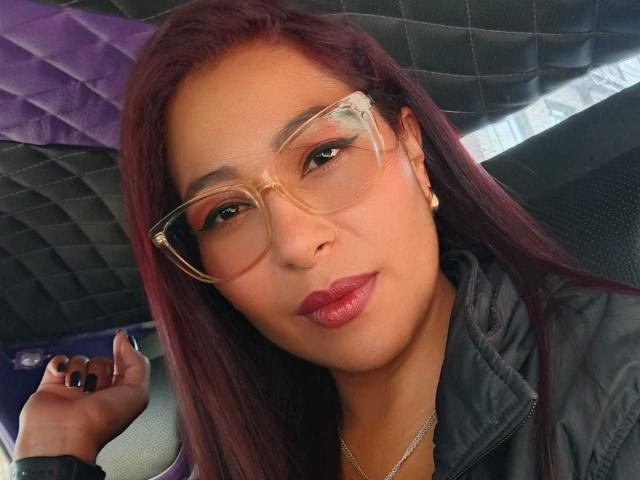 AishaFrench with long purple hair, clear glasses, and pink lipstick, wearing a gray jacket, holding a phone, and showing off her black nails. Her ass is not visible.
