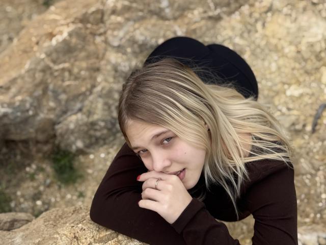 Belleza, con cabello rubio, camisa marrón de manga larga y pantalones negros, apoyada en las rocas, mordiéndose el dedo, mirando hacia arriba con una ligera sonrisa, mostrando su anillo..