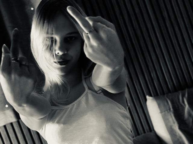 Photo en noir et blanc de Bellezzza, avec des cheveux blonds, portant un débardeur blanc, pointant avec ses deux doigts, montrant une bague à sa main droite, et ayant une expression sérieuse..