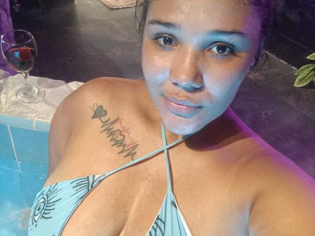 KennyRousse met natte huid, bruine ogen en een tatoeage op haar rechter schouder, draagt een blauw halterbikini, kijkt naar de camera en zit bij een zwembad met een drankje..
