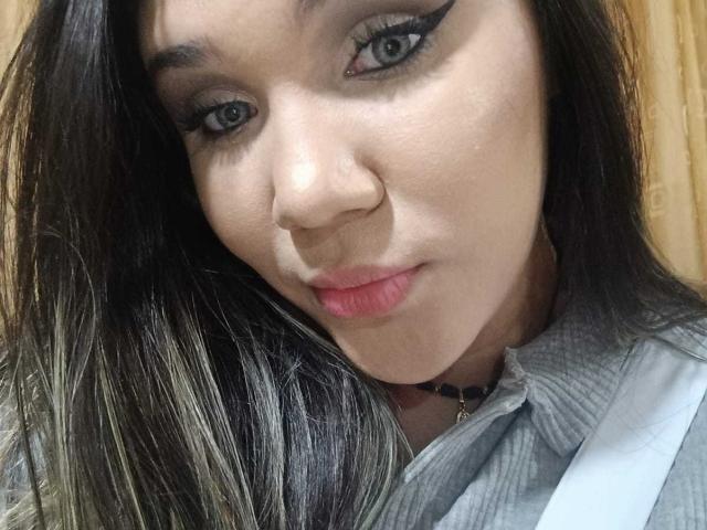 KennyRousse a une peau brun clair, un eye-liner noir, un rouge à lèvres rose et de longs cheveux foncés avec des mèches blondes. Elle porte une chemise grise avec un collier noir..