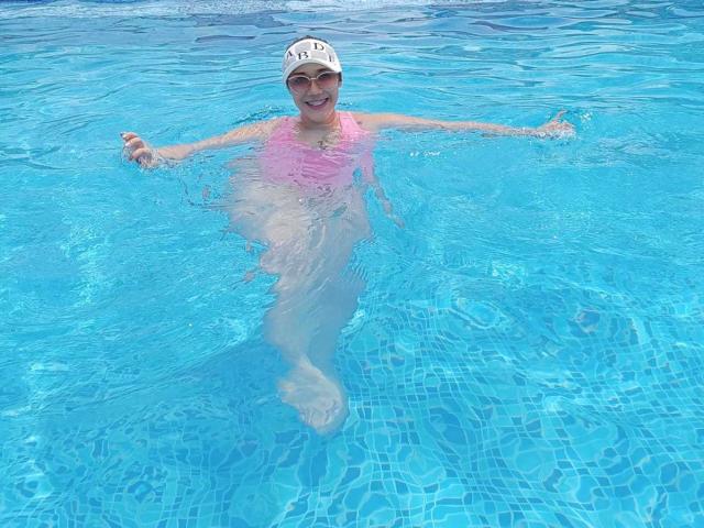 KennyRousse en una piscina azul, vistiendo un traje de baño rosa y un gorro blanco, con los brazos extendidos, sonriendo, con las piernas sumergidas, mostrando un trasero brillante y pequeños pechos firmes..