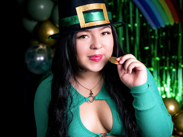 BarbaraHoneyy with long black hair, wearing a green top with cleavage, black top hat with gold buckle, eating a gold coin, pink lips, green background, rainbow.