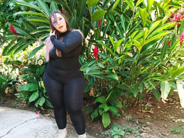 KennyRousse con un trasero grande, muslos gruesos, camiseta negra de manga larga, pantalones negros, cabello rubio y rosa, sombra de ojos azul, de pie frente a una planta verde con flores rojas..