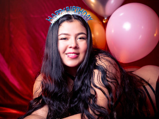 BarbaraHoneyy with long black wavy hair, wearing a "Happy Birthday" headband, smiling with pink lipstick, nose ring, and a lip piercing, surrounded by pink and gold balloons.