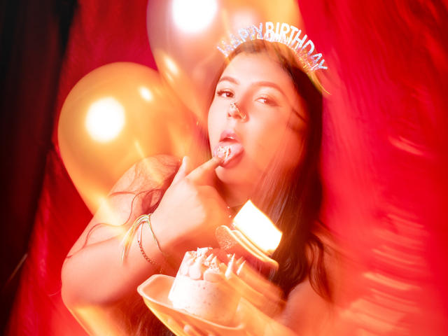 BarbaraHoneyy with a sweet tooth, licking frosting from a cake, wearing a "Happy Birthday" tiara, surrounded by gold balloons, holding a candle in her mouth.