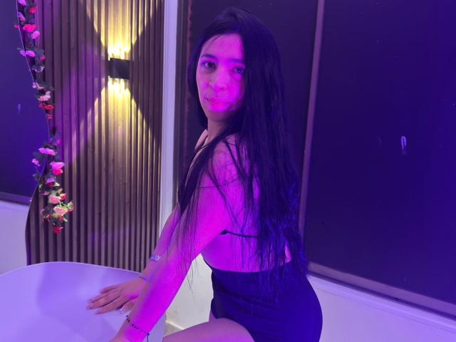 SamantaDiamont with long black hair, purple-lit, wearing a sheer black top, exposing her small tits, black shorts, and a bracelet, kneeling by a white chair, looking at camera.