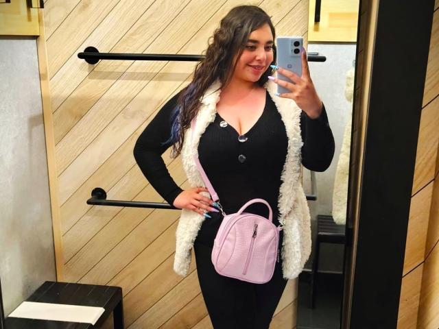 BaGiolett mit welligem, dunklem Haar, lächelnd, trägt ein schwarzes Langarmshirt, eine pinke Weste mit Knöpfen, eine weiße, fellartige Weste, eine pinke Handtasche, schwarze Hosen und hält ein Handy, in einem Selfie vor einem Spiegel..
