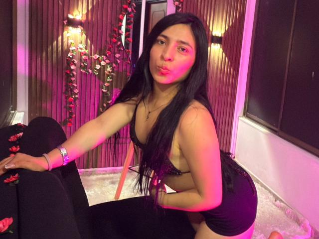SamantaDiamont, with long black hair, pouty lips, and a slim build, wearing a black lace bra and black skirt, kneeling on a bed with black leggings, and pink light.